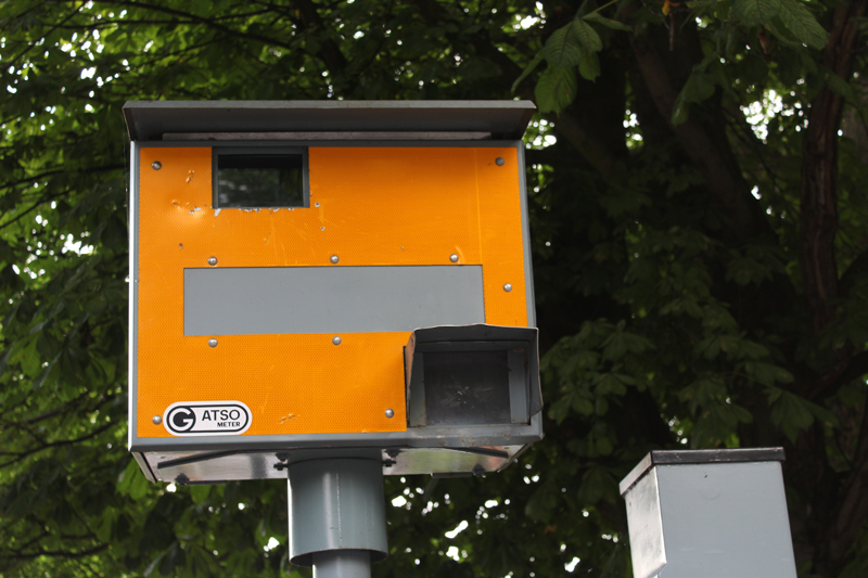 A yellow speed camera at a roadside with green leafy trees behind it