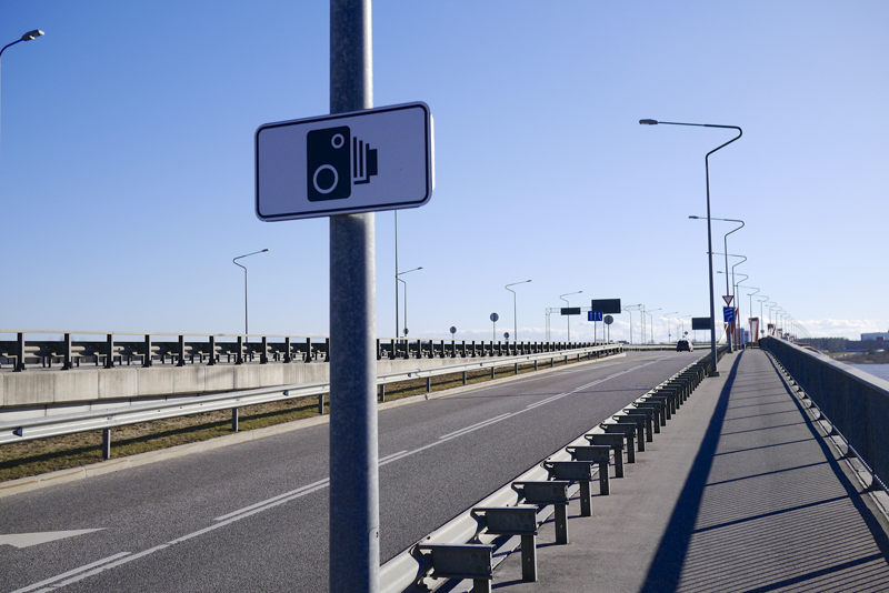A speed camera sign on pole next to slip road to a busy bridge